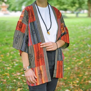 May include: A patchwork kimono-style jacket in shades of red, orange, black, and gray. The jacket features a striped pattern and decorative black designs. The sleeves are short, and the jacket is open in the front.