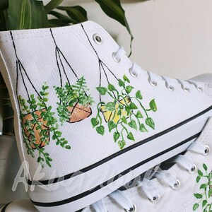 K&ouml;nnte beinhalten: Wei&szlig;e High-Top-Canvas-Schuhe mit handgemaltem Design von h&auml;ngenden Pflanzen in T&ouml;pfen. Die Pflanzen sind gr&uuml;n mit braunen und gelben T&ouml;pfen.
