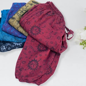 May include: A stack of five pairs of printed cotton trousers in various shades of blue, with a burgundy pair on top. The trousers have a sun and moon pattern.
