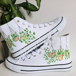 K&ouml;nnte beinhalten: Wei&szlig;e High-Top-Canvas-Schuhe mit handgemaltem Design von h&auml;ngenden Topfpflanzen. Die Pflanzen sind gr&uuml;n und gelb mit braunen T&ouml;pfen.