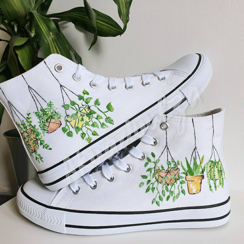 K&ouml;nnte beinhalten: Wei&szlig;e High-Top-Canvas-Schuhe mit handgemaltem Design von h&auml;ngenden Topfpflanzen. Die Pflanzen sind gr&uuml;n und gelb mit braunen T&ouml;pfen.
