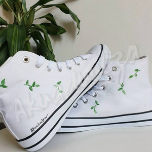 K&ouml;nnte beinhalten: Wei&szlig;e High-Top-Canvas-Sneaker mit gr&uuml;ner Blattembroidery und schwarzem Besatz. Die Schuhe haben eine handschriftliche Signatur, die "Akuatimba" lautet.