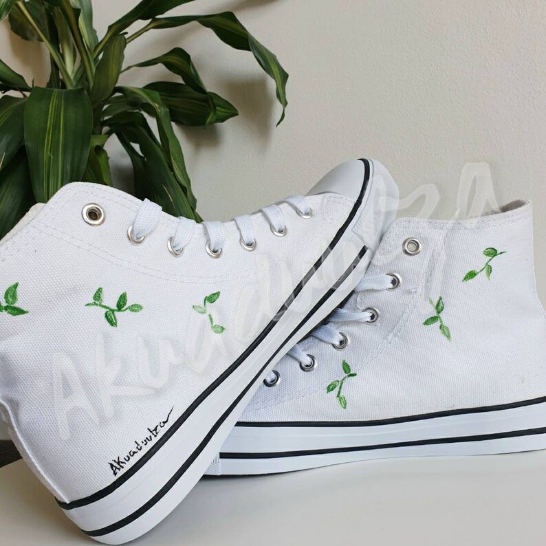 K&ouml;nnte beinhalten: Wei&szlig;e High-Top-Canvas-Sneaker mit gr&uuml;ner Blattembroidery und schwarzem Besatz. Die Schuhe haben eine handschriftliche Signatur, die "Akuatimba" lautet.