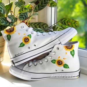 May include: White high-top canvas sneakers with hand-painted sunflowers. The sneakers have black trim and a black rubber sole.