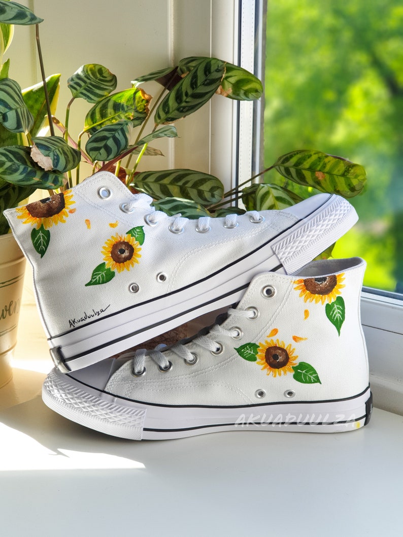 May include: White high-top canvas sneakers with hand-painted sunflowers. The sneakers have black trim and a black rubber sole.