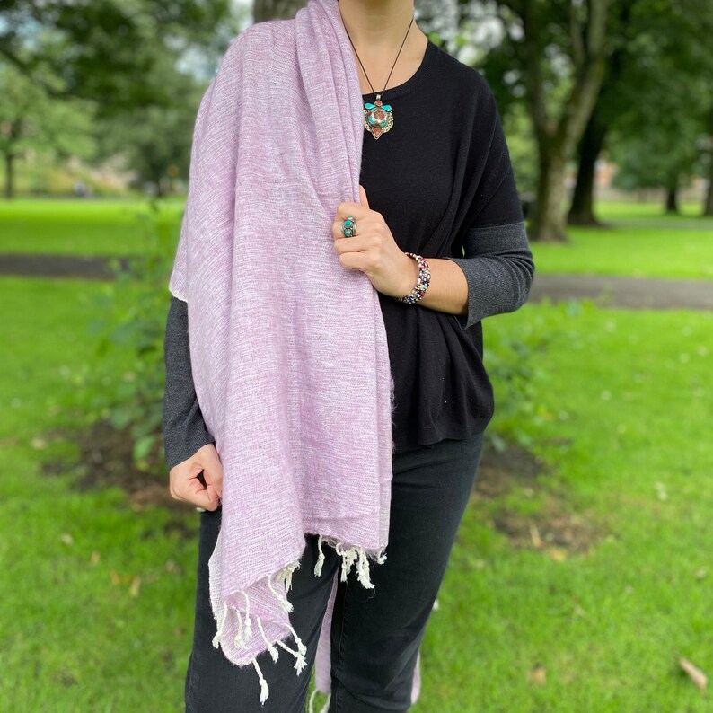 May include: A light purple pashmina scarf with white fringe. The scarf is draped around a person's shoulders and is worn with a black top and black pants.