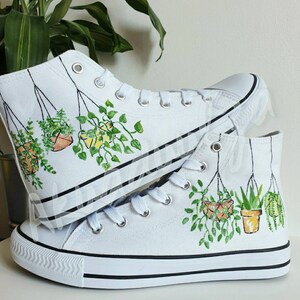 K&ouml;nnte beinhalten: Wei&szlig;e High-Top-Canvas-Schuhe mit handgemaltem Design von h&auml;ngenden Topfpflanzen. Die Pflanzen sind gr&uuml;n und die T&ouml;pfe sind orange und gelb.