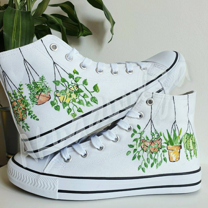 K&ouml;nnte beinhalten: Wei&szlig;e High-Top-Canvas-Schuhe mit handgemaltem Design von h&auml;ngenden Topfpflanzen. Die Pflanzen sind gr&uuml;n und die T&ouml;pfe sind orange und gelb.