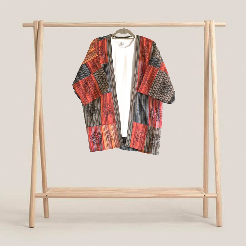 May include: A colorful patchwork kimono-style jacket with a white t-shirt underneath, hanging on a wooden clothing rack. The jacket features red, orange, black, and gray panels with embroidered floral designs. The rack is made of light-colored wood.