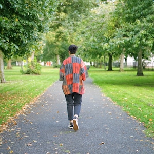 May include: A person walking away on a paved path in a park, wearing a patchwork jacket with red, orange, and gray panels. The jacket is paired with dark pants and sneakers. The scene is surrounded by green trees and grass.