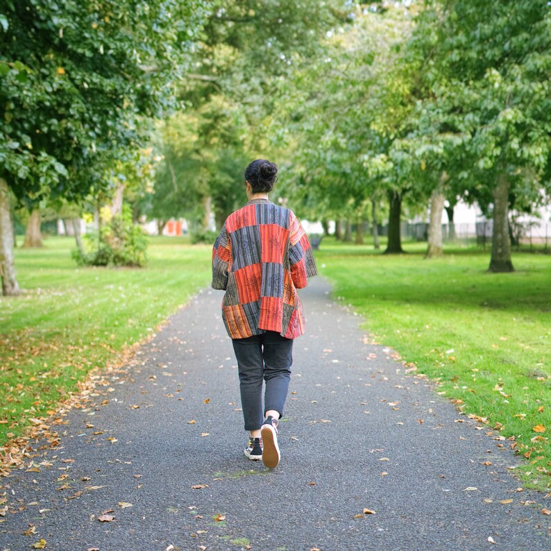 May include: A person walking away on a paved path in a park, wearing a patchwork jacket with red, orange, and gray panels. The jacket is paired with dark pants and sneakers. The scene is surrounded by green trees and grass.