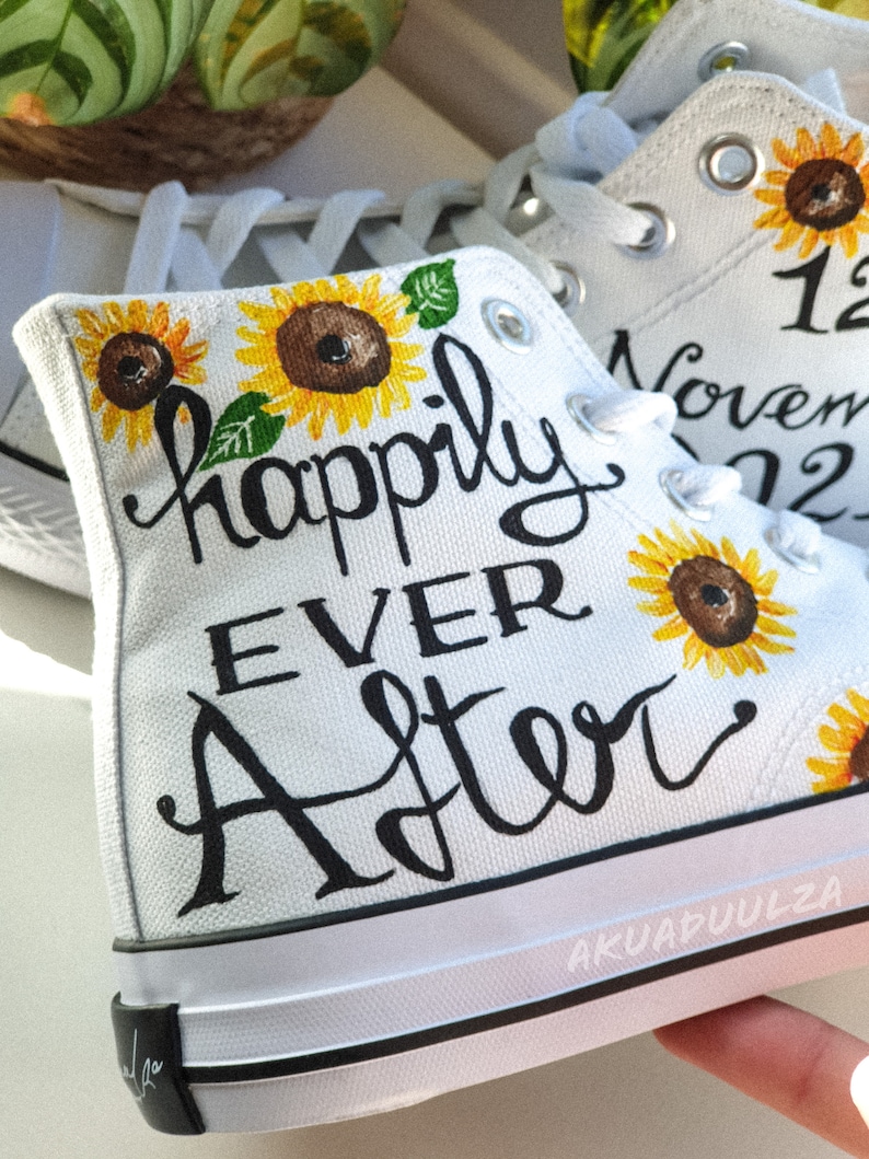 May include: White canvas high-top sneakers with black hand-painted lettering that reads "happily ever after". The sneakers are decorated with yellow sunflowers and the date "12 November 2022".
