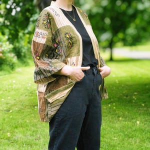 Peut inclure: Veste kimono patchwork aux tons verts, marron et beige. La veste pr&eacute;sente des motifs floraux brod&eacute;s et un devant ouvert. La personne porte un haut noir et un jean d&eacute;lav&eacute; fonc&eacute;. V&ecirc;tement d'ext&eacute;rieur &eacute;l&eacute;gant.