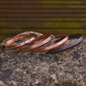 May include: Four copper bracelets with varying textures and designs are displayed on a gray stone. The bracelets have a warm, reddish-brown hue, with one appearing darker. The bracelets are of different widths and feature smooth, hammered, and patterned surfaces.