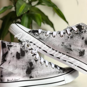 May include: White high-top canvas sneakers with black paint splatters and a black signature "Aknvas" on the side.