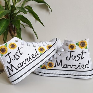 May include: A pair of white high-top canvas shoes with black laces and the words "Just Married" painted on them. The shoes are decorated with painted sunflowers.