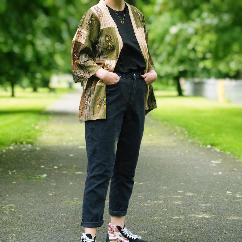 Peut inclure: Une personne porte une veste kimono &agrave; motifs avec un t-shirt noir et un jean d&eacute;lav&eacute; fonc&eacute;. La veste pr&eacute;sente un m&eacute;lange de panneaux verts, bruns et beiges avec des motifs floraux et g&eacute;om&eacute;triques. La tenue est compl&eacute;t&eacute;e par des baskets &agrave; motifs.