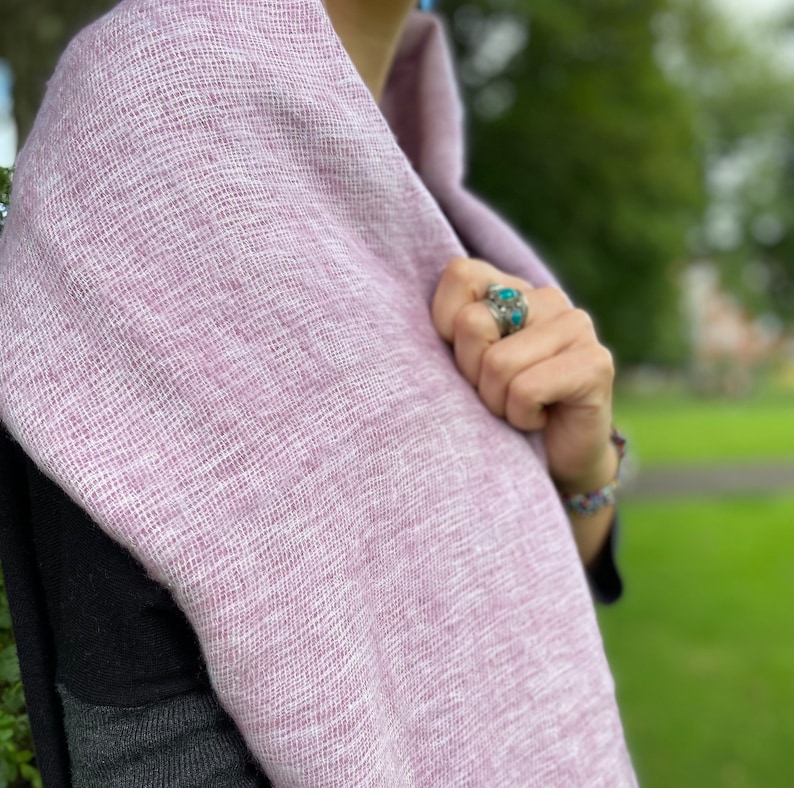 May include: A soft, light purple scarf made of a woven fabric. The scarf is draped around a person's neck and shoulders.