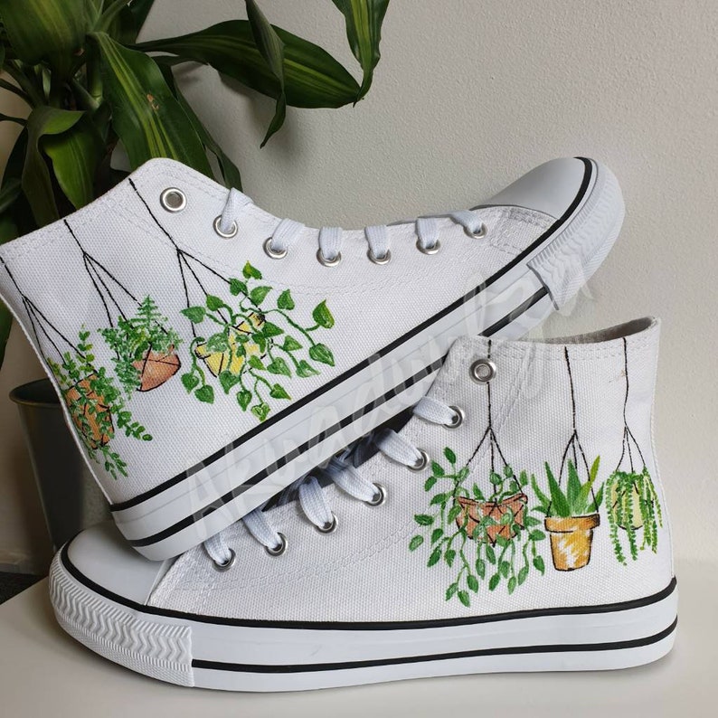 K&ouml;nnte beinhalten: Wei&szlig;e High-Top-Canvas-Schuhe mit handgemaltem Design von h&auml;ngenden Topfpflanzen. Die Pflanzen sind gr&uuml;n und braun mit einer Vielzahl von Blattformen.