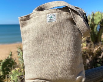 May include: A beige hemp tote bag with a herringbone weave pattern. The bag has a shoulder strap and a small label that reads "Himalayan Hemp THC Free". The bag is sitting on a wooden fence with a beach and blue sky in the background.