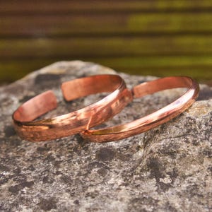 Op de afbeelding: Twee koperen manchetarmbanden met een gepolijste afwerking liggen op een grijs stenen oppervlak. De armbanden hebben een eenvoudig, open ontwerp. Het koper heeft een warme, roodbruine tint die licht reflecteert. De armbanden zijn waarschijnlijk handgemaakt.