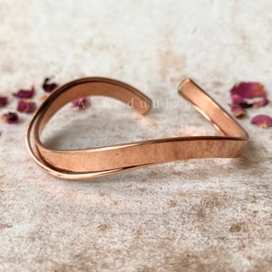 May include: A simple copper bracelet with a wavy design. The bracelet is made of two pieces of copper wire that are twisted together.