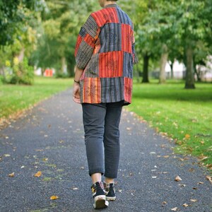 May include: A person wearing a patchwork kimono-style shirt with red, orange, and blue panels, paired with dark gray pants and black sneakers. The shirt features floral embroidery. The person is walking on a paved path.