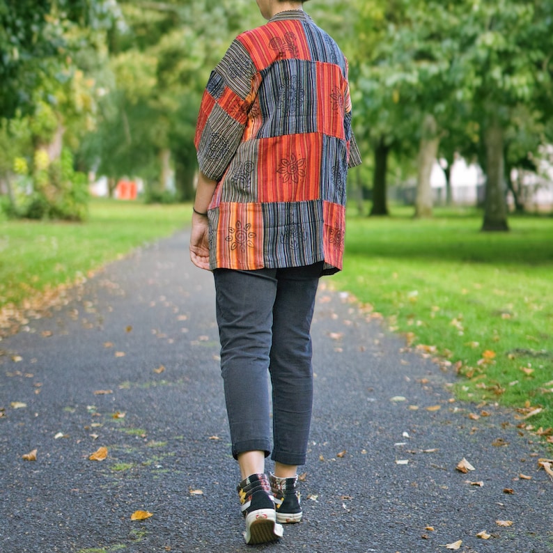 May include: A person wearing a patchwork kimono-style shirt with red, orange, and blue panels, paired with dark gray pants and black sneakers. The shirt features floral embroidery. The person is walking on a paved path.