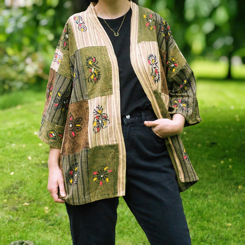 Peut inclure: Veste kimono patchwork aux tons verts, marron et beige. La veste pr&eacute;sente des motifs floraux et g&eacute;om&eacute;triques, avec des manches trois-quarts. Le v&ecirc;tement est port&eacute; sur un haut noir et un jean noir.