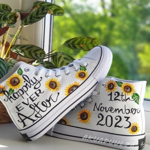 May include: A pair of white canvas high-top sneakers with black trim. The sneakers are decorated with hand-painted sunflowers and the text "happily ever after" on one shoe and "12th November 2023" on the other.