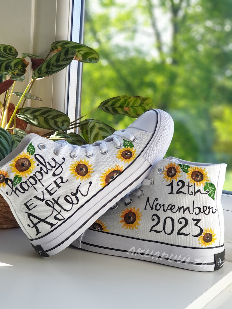 May include: A pair of white canvas high-top sneakers with black trim. The sneakers are decorated with hand-painted sunflowers and the text "happily ever after" on one shoe and "12th November 2023" on the other.