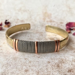 Könnte beinhalten: Ein Messing-Armreif mit einem geflochtenen Kupferband. Das Armband ist schlicht und elegant mit einem dezenten Design.