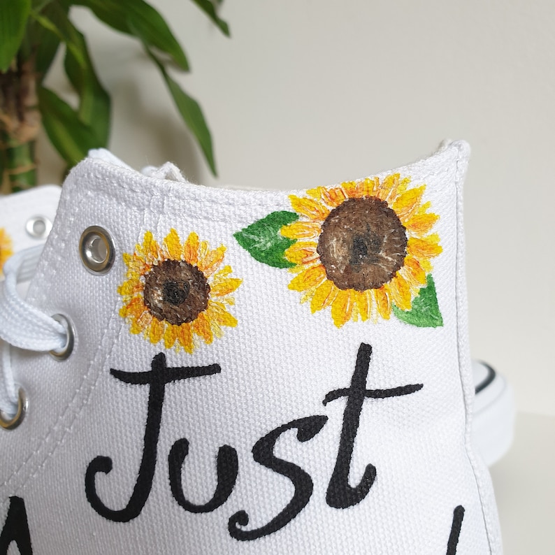May include: White canvas high-top sneakers with a hand-painted design of sunflowers and the word "Just" in black script.