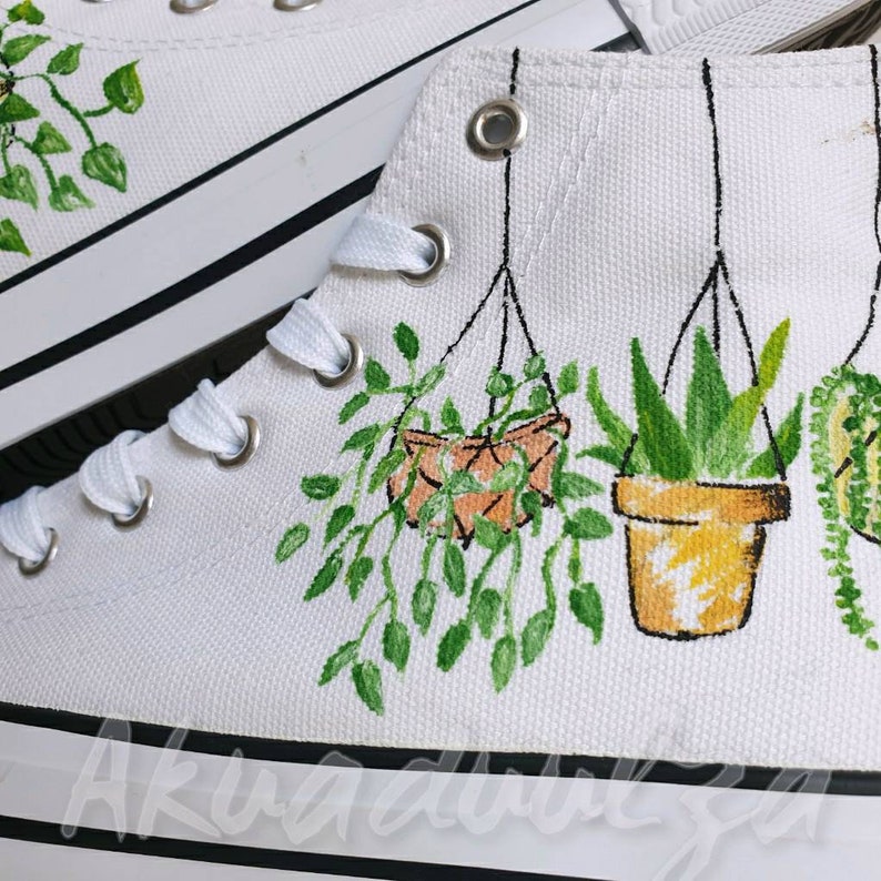 K&ouml;nnte beinhalten: Wei&szlig;e Canvas-High-Top-Sneaker mit handgemaltem Design von h&auml;ngenden Topfpflanzen. Die Pflanzen sind gr&uuml;n und braun mit gelben Akzenten. Die Sneaker haben schwarze Schn&uuml;rsenkel und einen schwarzen Streifen um die Sohle.