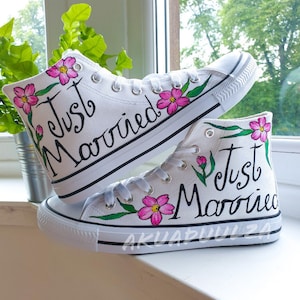 Könnte beinhalten: Ein Paar weiße High-Top-Canvas-Schuhe mit pinkfarbenen Blumen und grünen Blättern, die darauf gemalt sind. Die Schuhe sagen "Just Married" in schwarzer Schrift.