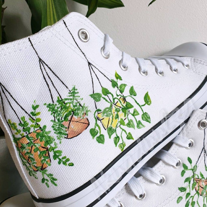 K&ouml;nnte beinhalten: Wei&szlig;e High-Top-Canvas-Schuhe mit handgemaltem Design von h&auml;ngenden Pflanzen. Die Pflanzen sind gr&uuml;n mit braunen T&ouml;pfen und in einem realistischen Stil gemalt.