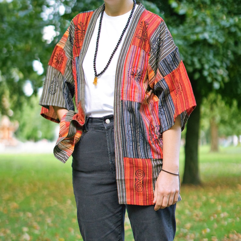 May include: A patchwork kimono-style jacket with a vibrant mix of colors including red, orange, black, and gray. The jacket features a variety of patterns and designs, and is worn over a white t-shirt and black jeans. A beaded necklace is also visible.
