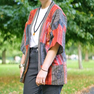 May include: A colorful patchwork kimono-style jacket with vertical stripes in red, orange, black, and blue. The jacket has short sleeves and a loose fit. The person is wearing a white t-shirt and black jeans. A long beaded necklace and bracelets complete the look.