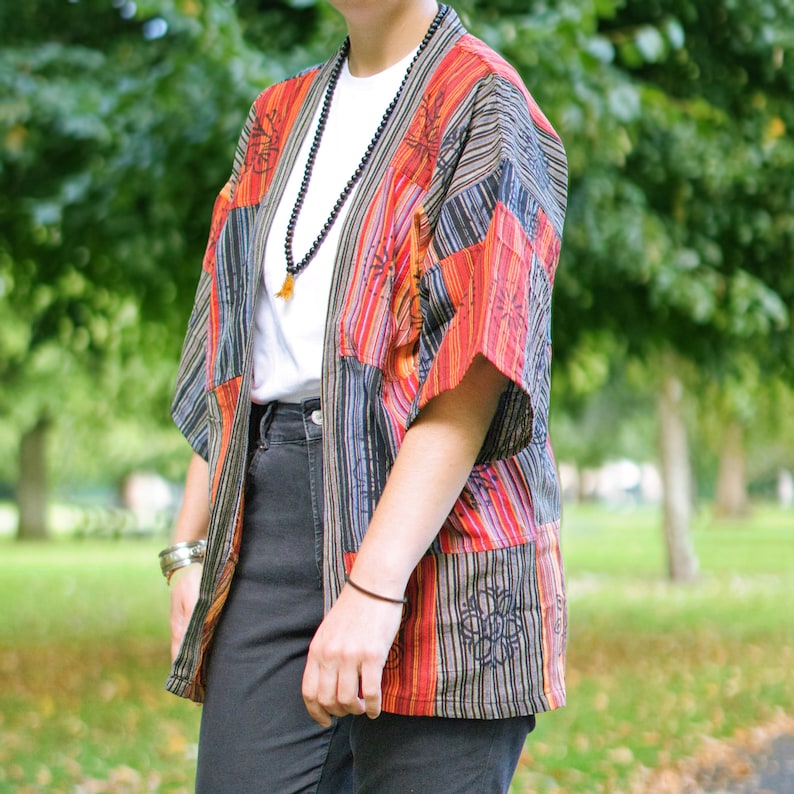 May include: A colorful patchwork kimono-style jacket with vertical stripes in red, orange, black, and blue. The jacket has short sleeves and a loose fit. The person is wearing a white t-shirt and black jeans. A long beaded necklace and bracelets complete the look.