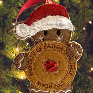 May include: A festive Christmas tree ornament in the shape of a gingerbread man, wearing a red and white Santa hat. The ornament has a gold-coloured disc that reads "PROPERTY OF FATHER CHRISTMAS NORTH POLE."