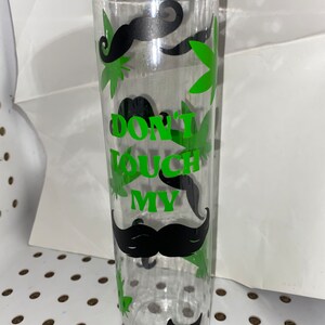 Puede incluir: Botella de agua de plástico transparente con tapa negra. La botella está decorada con hojas de cannabis verdes, bigotes negros y el texto "DON'T TOUCH MY" en verde.