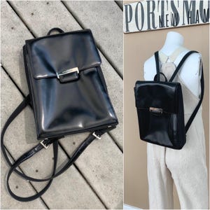 May include: A black leather backpack with a rectangular shape and a flap closure. The backpack features adjustable straps and a silver-colored metal clasp. The bag is displayed on a wooden surface and on a mannequin.