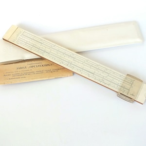 May include: A vintage slide rule with a white plastic case. The slide rule is made of wood and has a white face with black markings. The case is labelled "Завод "ОРГАТЕХНИКА" in Cyrillic lettering.