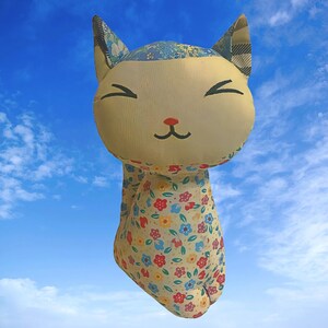 以下が含まれることがあります： 笑顔の顔と花柄のぬいぐるみ猫のおもちゃ。猫は薄い黄色で、ピンクの鼻と青い目。花柄はピンク、ブルー、イエロー、グリーンの色合いです。