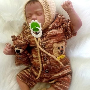 Könnte beinhalten: Eine gestrickte Baby-Puppe, die ein gestricktes Outfit in Braun- und Beigetönen mit einem Bären-Applikationen trägt. Die Puppe trägt eine gestrickte Mütze und hat einen Schnuller im Mund.