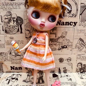 May include: A doll with blonde hair and blue eyes wearing a striped orange and white dress and white shoes. The doll is holding a small ice cream cone. The doll is standing in front of a background of colorful patterned paper with the word "Nancy" written on it.