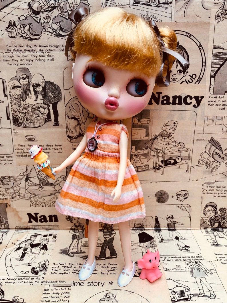 May include: A doll with blonde hair and blue eyes wearing a striped orange and white dress and white shoes. The doll is holding a small ice cream cone. The doll is standing in front of a background of colorful patterned paper with the word "Nancy" written on it.