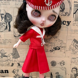 Puede incluir: Mu&ntilde;eca con un atuendo de marinero rojo, que incluye boina con emblema de ancla, chaqueta y falda plisada. La mu&ntilde;eca tiene cabello oscuro, piel p&aacute;lida y botas negras. El fondo es un peri&oacute;dico vintage.