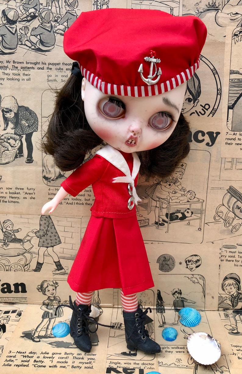 Puede incluir: Mu&ntilde;eca con un atuendo de marinero rojo, que incluye boina con emblema de ancla, chaqueta y falda plisada. La mu&ntilde;eca tiene cabello oscuro, piel p&aacute;lida y botas negras. El fondo es un peri&oacute;dico vintage.