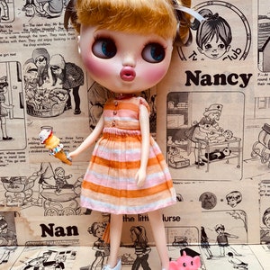 May include: A doll with blonde hair and blue eyes wearing a pink and orange striped dress and white shoes. The doll is holding a yellow and orange bird toy. The doll is standing in front of a vintage magazine page with the text "Nancy" and "the little nurse".